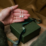 Hand holding pearl earrings above a green jewelry box on a patterned surface