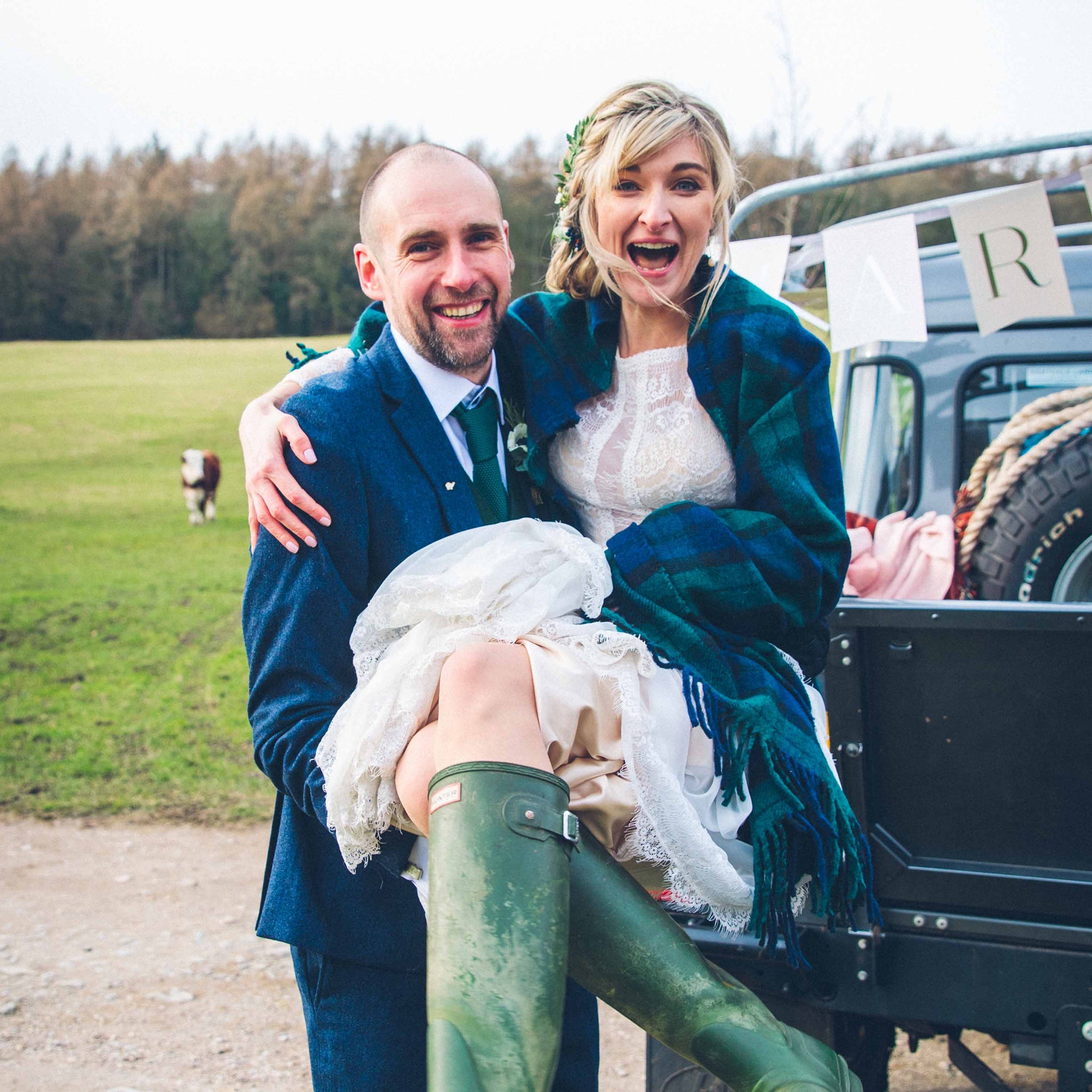 husband carrying wife. She is wearing a wedding dress and green wellington boots.